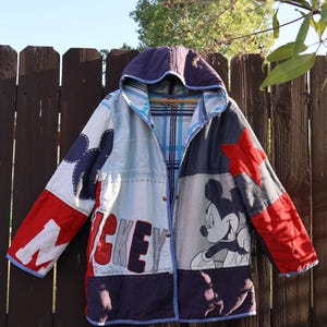 Puede incluir: Una chaqueta con capucha de diseño patchwork con paneles en rojo, blanco, azul y morado. La prenda muestra un gráfico de Mickey Mouse, el texto "MICKEY" en letras grandes y una aplicación de estrella roja. Tiene cierre frontal de botones.