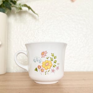 May include: White ceramic mug with a floral design featuring yellow, pink, and blue flowers. The mug has a curved handle and a slightly flared rim. The flowers are surrounded by green leaves and stems.
