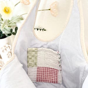 May include: A light blue tote bag with a cream-coloured trim and a square, quilted patch pocket on the interior. The pocket features a patchwork design with sections of green plaid, white fabric, and red gingham, finished with visible stitching.