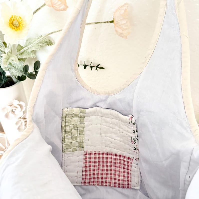 May include: A light blue tote bag with a cream-coloured trim and a square, quilted patch pocket on the interior. The pocket features a patchwork design with sections of green plaid, white fabric, and red gingham, finished with visible stitching.