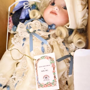 May include: A porcelain doll with blonde curls wearing a cream-coloured dress with lace trim, blue ribbons, and a matching bonnet. The doll features blue eyes and a tag reading American Artists Collection by KAIS, INC. and World Gallery Dolls.