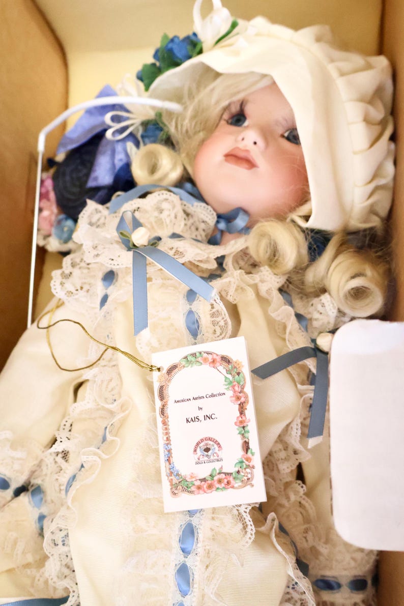 May include: A porcelain doll with blonde curls wearing a cream-coloured dress with lace trim, blue ribbons, and a matching bonnet. The doll features blue eyes and a tag reading American Artists Collection by KAIS, INC. and World Gallery Dolls.