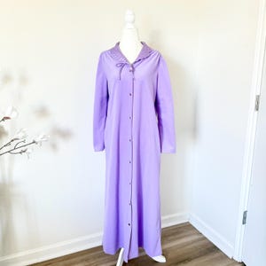 Puede incluir: Una bata larga de color lavanda con mangas largas y botones en la parte delantera. La bata presenta un cuello decorativo con borde festoneado y un cierre de lazo en el escote. La bata se exhibe en un maniquí blanco.