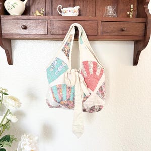 May include: A quilted hobo bag featuring a fan-patterned patchwork design in shades of coral, teal, and floral prints. The bag includes a long, solid cream-coloured fabric tie closure at the centre and a single integrated shoulder strap.