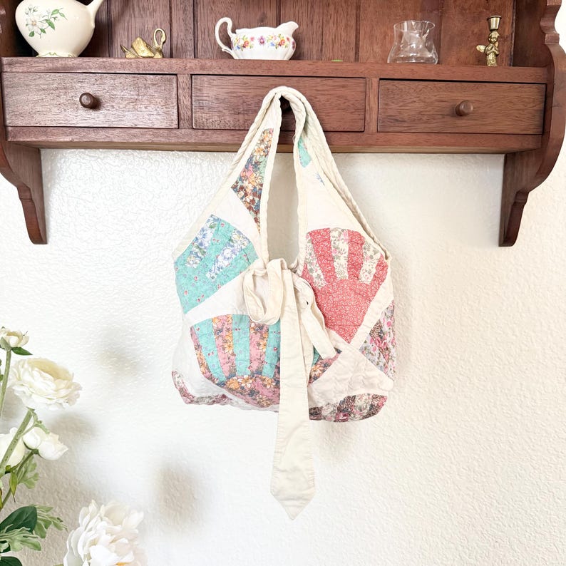 May include: A quilted hobo bag featuring a fan-patterned patchwork design in shades of coral, teal, and floral prints. The bag includes a long, solid cream-coloured fabric tie closure at the centre and a single integrated shoulder strap.