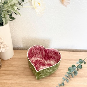 Puede incluir: Un cuenco decorativo en forma de corazón con interior rojo y exterior verde. El cuenco tiene una superficie texturizada y un borde blanco. El cuenco está sobre una superficie de madera.