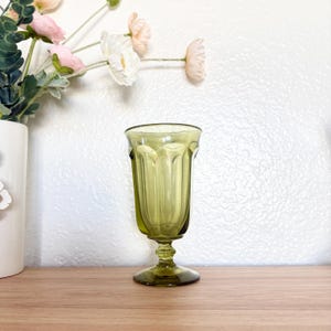 May include: An olive green vintage-style glass with a pedestal base. The glass has a fluted design and a wide rim. The background includes a white wall and a wooden surface. Artificial flowers in a white vase are visible on the left.