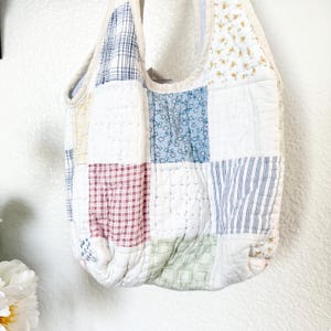 May include: A quilted hobo bag constructed from various fabric squares featuring a mix of patterns including blue floral, red and white gingham, green plaid, blue stripes, and yellow floral prints, all stitched together with a white fabric trim.