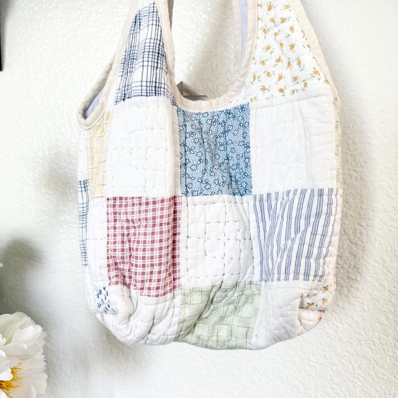 May include: A quilted hobo bag constructed from various fabric squares featuring a mix of patterns including blue floral, red and white gingham, green plaid, blue stripes, and yellow floral prints, all stitched together with a white fabric trim.