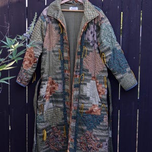 May include: A long, quilted coat featuring a patchwork design of various earth-toned fabrics in shades of olive green, brown, and tan. The garment includes a wide collar, long sleeves, and yellow cord ties along the front opening and cuffs.
