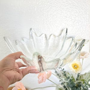 Può includere: Una ciotola di vetro trasparente con un design unico a forma di petalo. La ciotola è tenuta in mano, mostrando la sua forma e consistenza. I bordi della ciotola hanno un design ondulato e appuntito. Lo sfondo include fiori bianchi e rosa.