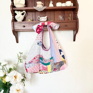 May include: A hobo-style shoulder bag constructed from a patchwork of various fabrics including floral, gingham, striped, and paisley patterns in shades of pink, blue, green, yellow, and white, featuring a large fabric bow at the top strap.