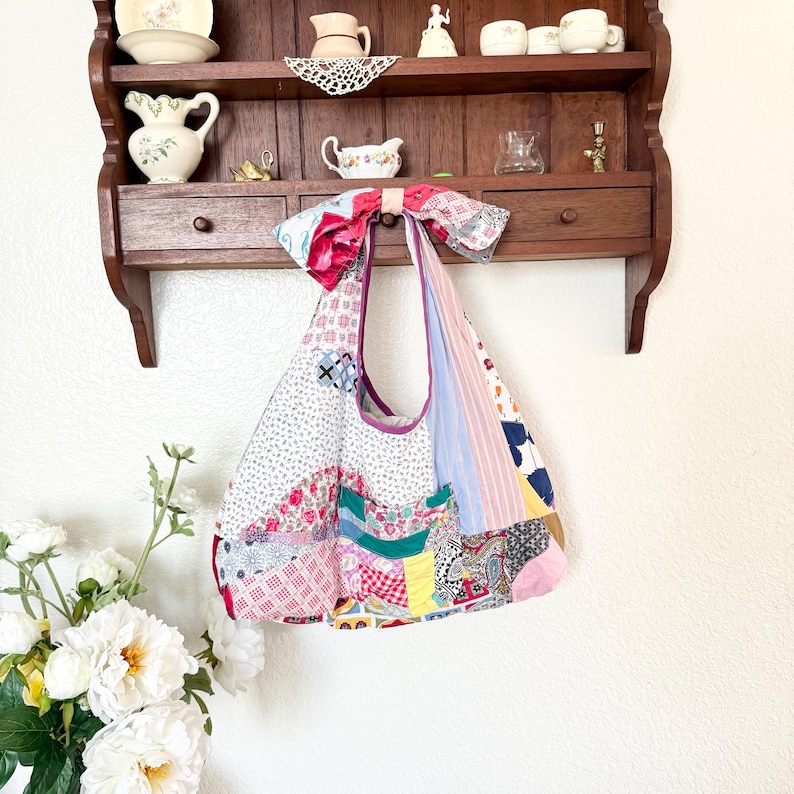 May include: A hobo-style shoulder bag constructed from a patchwork of various fabrics including floral, gingham, striped, and paisley patterns in shades of pink, blue, green, yellow, and white, featuring a large fabric bow at the top strap.