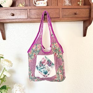 May include: A quilted tote bag featuring a patchwork design with floral patterns in shades of pink, green, and white. The bag has a central white square panel with a diamond-shaped floral print, purple fabric trim, and two matching purple shoulder straps.