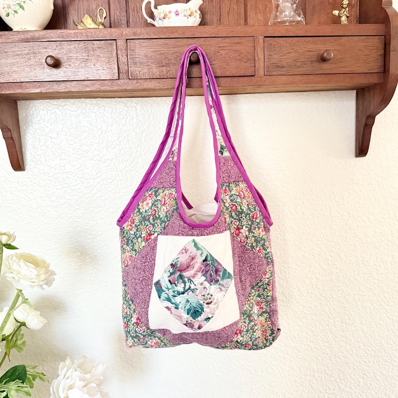 May include: A quilted tote bag featuring a patchwork design with floral patterns in shades of pink, green, and white. The bag has a central white square panel with a diamond-shaped floral print, purple fabric trim, and two matching purple shoulder straps.
