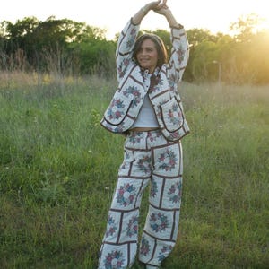May include: A quilted jacket and matching wide-leg trousers set featuring a white base with a repeating floral star pattern in shades of blue, red, and green, accented by brown trim. The set is worn over a plain white cropped tank top.