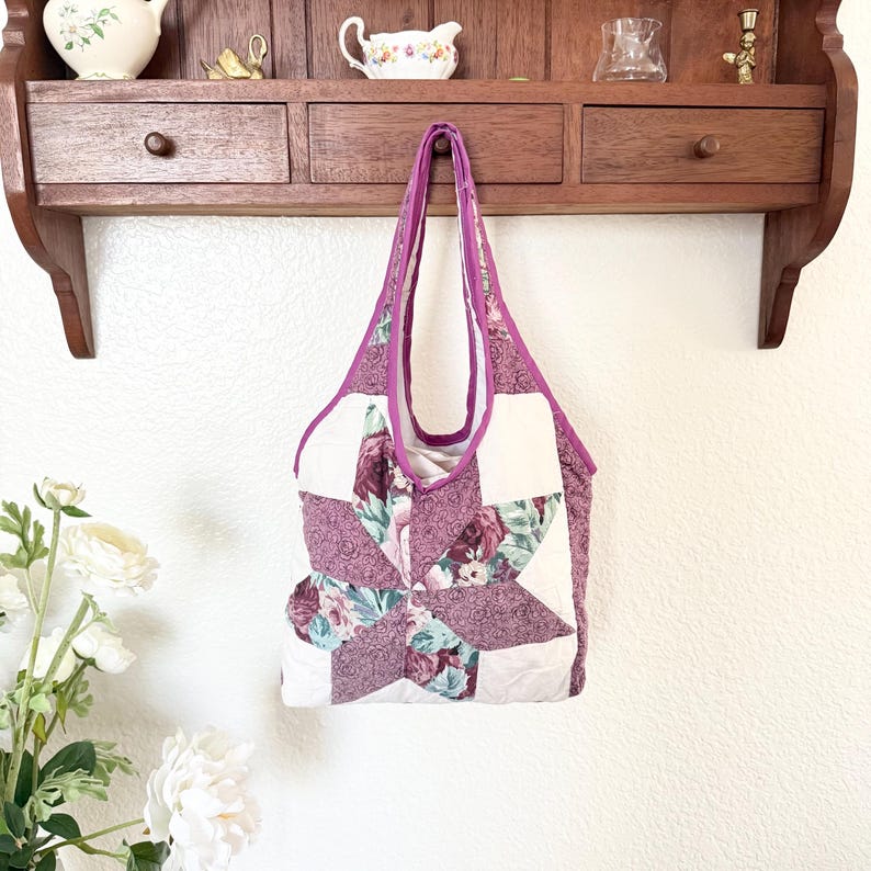 May include: A quilted tote bag featuring a star-shaped patchwork design in shades of mauve, dusty rose, and cream. The bag has a floral pattern on the star points, solid cream sections, and purple fabric trim along the edges and the shoulder straps.