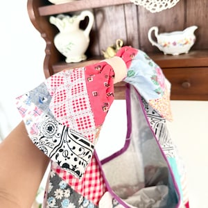 May include: A patchwork tote bag constructed from various fabric scraps featuring red and white gingham, black and white paisley, floral patterns, and solid pink sections. The bag has a knotted handle and a purple trim along the interior opening.