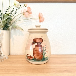 May include: A beige ceramic jar with a lid, featuring a hand-painted scene of a well, a basket, and flowers. The jar is on a wooden surface, with flowers in a vase in the background.