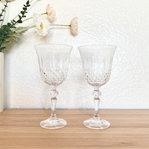 May include: Two clear crystal wine glasses with diamond-cut patterns. The glasses have a flared bowl, a decorative stem, and a round base. The glasses are displayed on a wooden surface with white flowers in the background.