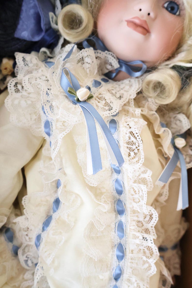 May include: A porcelain doll with blonde hair and blue eyes, wearing a cream-coloured dress adorned with intricate white lace ruffles, light blue satin ribbon trim, and small decorative white rosebuds at the neckline and sleeves.