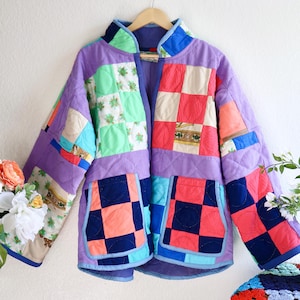 May include: A quilted jacket featuring a patchwork design of various fabric squares in shades of purple, red, light blue, navy, and green. The garment includes a stand-up collar, long sleeves, and two large front patch pockets with a checkered pattern.