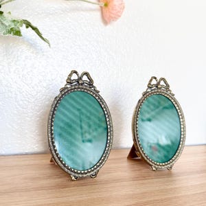 May include: Two antique oval picture frames with a bow detail at the top. The frames have a bronze finish and a green background. They are standing on a wooden surface.