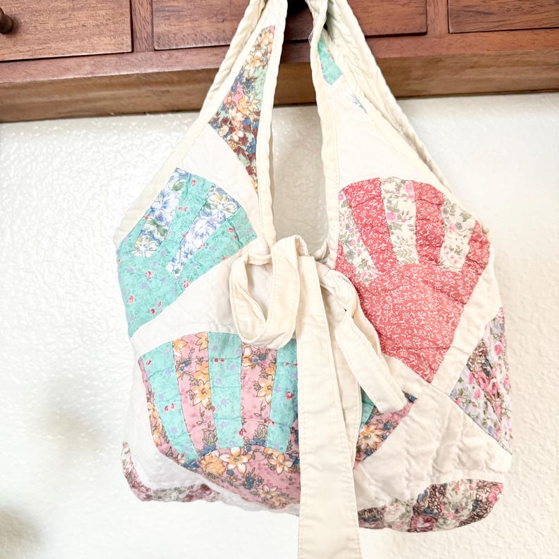 May include: A quilted hobo bag featuring a fan-shaped patchwork design in pastel floral patterns of pink, green, and red on a cream background. The bag includes a long, cream-coloured fabric tie closure and a wide, integrated shoulder strap.
