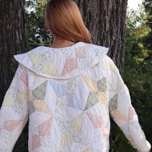 May include: A long-sleeved quilted jacket featuring a patchwork design of white, pale yellow, light blue, and soft pink fabric segments. The garment includes a wide, folded collar and a repeating geometric star pattern across the back and sleeves.