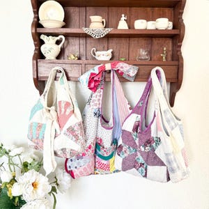 May include: Three handmade hobo-style bags crafted from repurposed vintage quilt fabric. The bags feature various patchwork patterns in floral, plaid, and geometric designs with colours including pink, purple, green, and white. Each bag has a wide shoulder strap.