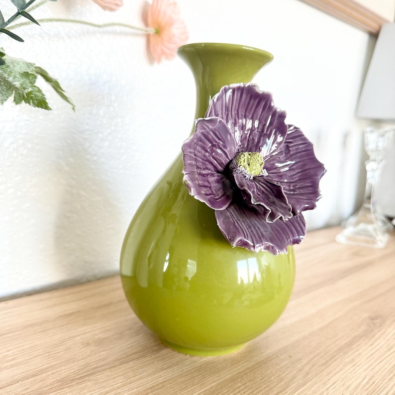 May include: A glossy, green ceramic vase with a teardrop shape and a flared top. A large, textured purple flower with a yellow center is attached to the vase. The vase is displayed on a wooden surface.