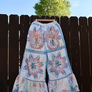 Puede incluir: Pantalones de pierna ancha confeccionados en tela acolchada con un diseño de patchwork de patrones florales en rosa y azul. Los pantalones incluyen dos grandes bolsillos delanteros con lazos de cinta rosa y un dobladillo con volantes.