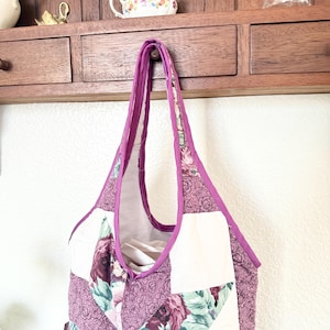 May include: A quilted tote bag featuring a patchwork design of solid white fabric and a floral pattern in shades of purple, mauve, and green. The bag has a deep purple fabric trim along the edges and two integrated shoulder straps.