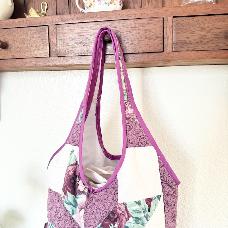 May include: A quilted tote bag featuring a patchwork design of solid white fabric and a floral pattern in shades of purple, mauve, and green. The bag has a deep purple fabric trim along the edges and two integrated shoulder straps.