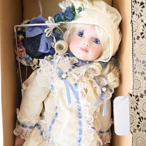 May include: A porcelain doll with blonde hair and blue eyes, wearing a cream-coloured dress with lace trim and blue ribbon accents. The doll wears a matching cream bonnet adorned with blue flowers and is positioned inside a cardboard box.