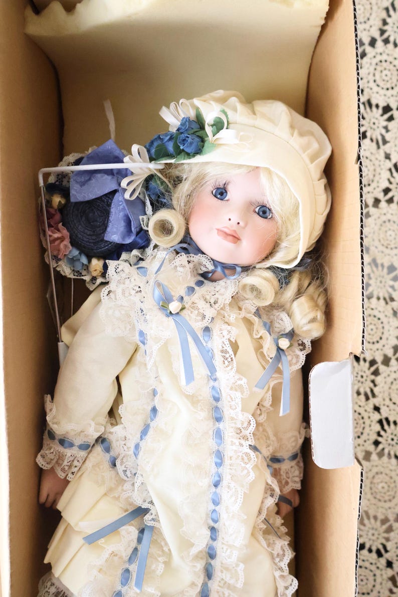 May include: A porcelain doll with blonde hair and blue eyes, wearing a cream-coloured dress with lace trim and blue ribbon accents. The doll wears a matching cream bonnet adorned with blue flowers and is positioned inside a cardboard box.