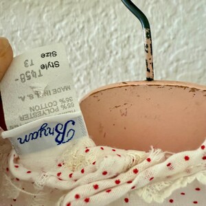 May include: Close-up of a white and red polka dot garment with a lace trim, hanging on a vintage pink hanger. The tag reads "Bryan" and "Made in U.S.A." with the style and size information.