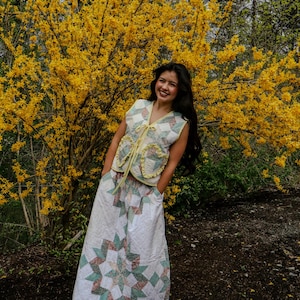 May include: A quilted vest and skirt set in a patchwork design. The vest has a V-neck and ruffled pockets, with a yellow tie. The skirt features a large star pattern. The outfit is in shades of white, green, and pink.