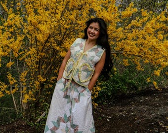 May include: A patchwork quilted vest and skirt. The vest has a V-neck and ruffled pockets, with a yellow tie. The skirt features a large star pattern. The outfit is in white, green, and pink. The outfit is handmade.