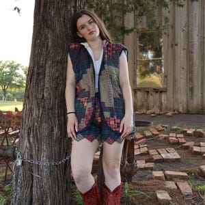 May include: A quilted vest and shorts set in a patchwork design with shades of green, red, and brown. The outfit is paired with red cowboy boots. The model is leaning against a tree in front of a wooden building.