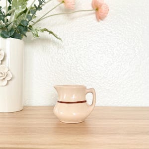 May include: A small, cream-colored ceramic creamer with a dark brown stripe. The creamer has a handle and a spout. A white vase with floral accents and pink flowers are in the background. The creamer is on a wooden surface.