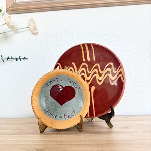 May include: Two round ceramic plates displayed on metal stands. The smaller front plate is light blue with a dark red heart and the text "EAST or WEST Home is Best". The larger back plate is dark red with yellow wavy lines and vertical stripes.