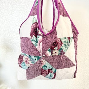 May include: A quilted tote bag featuring a star-shaped pattern made from fabric with a floral print in shades of mauve, dark red, and teal, contrasted with solid white fabric panels and finished with a solid mauve fabric trim along the edges.