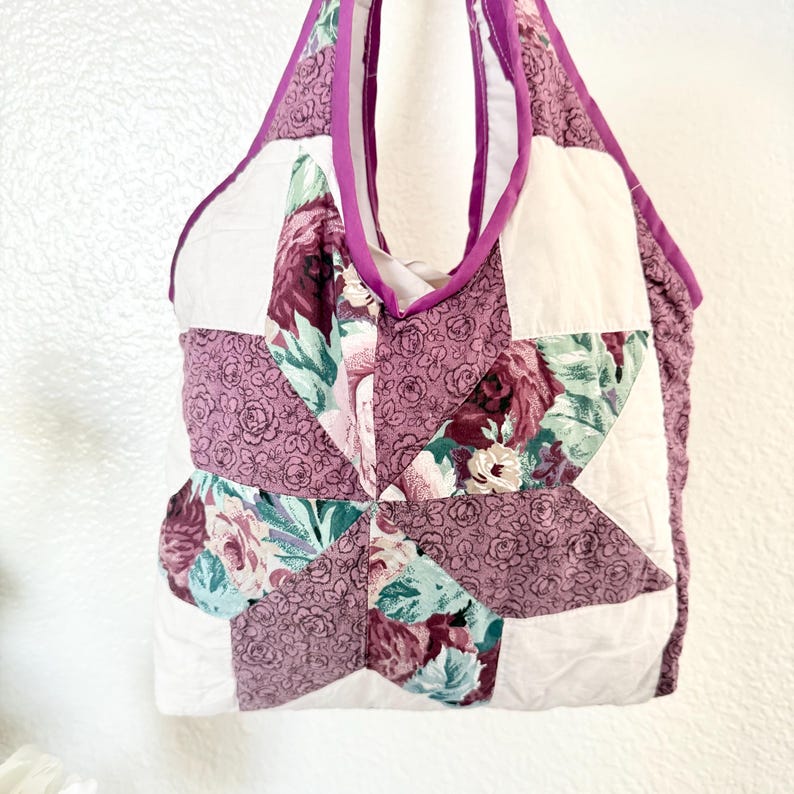 May include: A quilted tote bag featuring a star-shaped pattern made from fabric with a floral print in shades of mauve, dark red, and teal, contrasted with solid white fabric panels and finished with a solid mauve fabric trim along the edges.