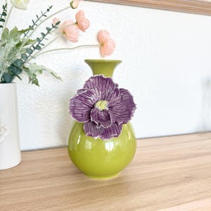 May include: A green ceramic vase with a large, purple flower accent. The flower has a yellow center. The vase is round and has a narrow neck. The vase is sitting on a wooden surface.