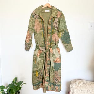 May include: A long patchwork robe in shades of green, tan, and brown hangs on a wooden hanger. The robe features a collared neckline, long sleeves, and a matching belt. Yellow tassels accent the front. The robe is made of various patterned fabrics.