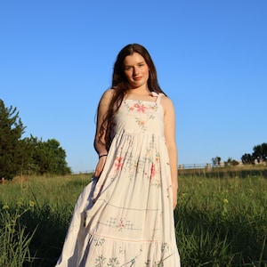 Puede incluir: Un vestido de verano blanco con bordados florales y falda escalonada. El vestido presenta detalles florales rosas y verdes y un escote cuadrado. El vestido se usa al aire libre en un campo de hierba alta bajo un cielo azul.