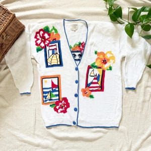 May include: A white long-sleeve V-neck cardigan sweater featuring a blue trim and a series of colorful knit patterns. The designs include rectangular frames with sailboats, beach umbrellas, and vibrant red, orange, and yellow floral motifs.