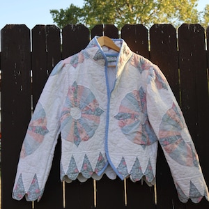 May include: A quilted jacket featuring a white base with circular fan-shaped patterns in pastel pink, blue, and floral prints. The garment has a high collar, long sleeves, a light blue trim along the edges, and a scalloped hemline with triangular accents.