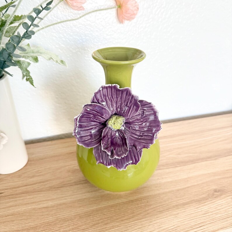 May include: A ceramic vase in shades of green and purple. The vase has a round base and a slender neck. A large, textured purple flower with a yellow center is attached to the vase. The vase is a decorative home accent.